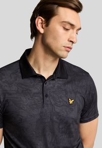 Sort polo shirt med tekstureret mønster, udstyret med en slank krave og et guld logo på venstre bryst. Korte ærmer og knaplukning.