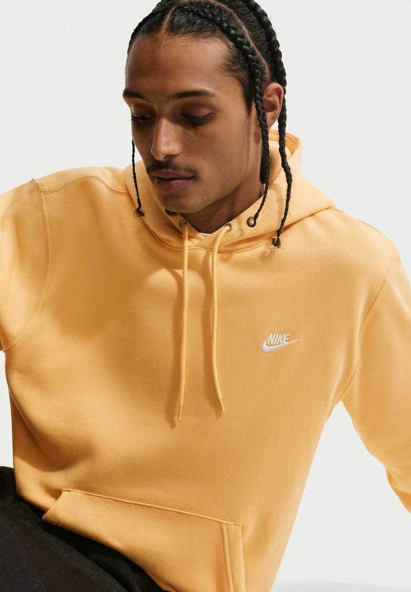 Junger Mann mit geflochtenem Haar, der einen gelben Nike-Hoodie trägt und nach unten schaut vor einem schlichten hellen Hintergrund.