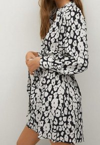 Mujer ajustando el cinturón en un vestido floral en blanco y negro con mangas largas y un dobladillo corto, frente a un fondo liso.