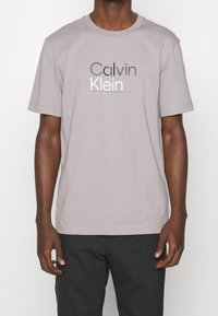 Grå bomulds t-shirt med "Calvin Klein" logo i kontrasterende sort og hvid tekst. Korte ærmer og standard crew neck design.