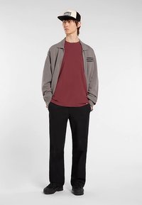 Giacca grigia con zip, t-shirt marrone scuro, pantaloni neri e scarpe nere. Il cappellino ha una parte frontale color crema con dettagli neri. Stile semplice e casual.