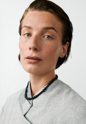 Persoană cu păr scurt, brunet, pieptănat pe spate, purtând haine gri cu textură și un colier negru cu mărgele, pe un fundal alb simplu.