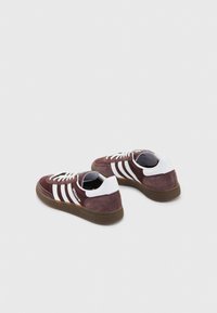 Sneakers in camoscio marrone con dettagli bianchi, caratterizzate da tre strisce bianche, suole in gomma e design classico con lacci. Sul retro è presente il dettaglio del marchio.