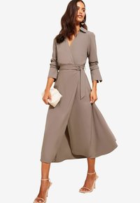 Langes, beiges Wickelkleid mit Kragen und tailliertem Bund. Mit Dreiviertelärmeln und einem fließenden Rock. Stylisch kombiniert mit Riemchen-Heels und einer Clutch.