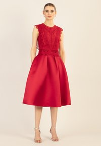 Rotes Kleid mit Spitzenoberteil, gewelltem Saum, rundem Ausschnitt und satinierter A-Linien-Rock. Silberne Sandalen ergänzen das Outfit.