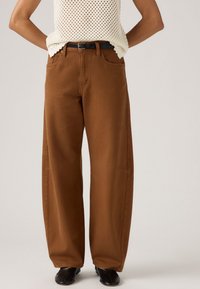 Persoon in hooggeplooide, wijde bruine broek met een zwarte riem, een crèmekleurige haakjesmouwloze top en zwarte vierkante schoenen.