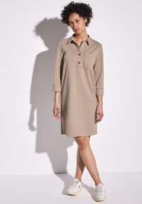 Beige jurk met kraag, knoopsluiting, driekwart mouwen en een borstzak, gecombineerd met witte sneakers op een neutrale achtergrond.