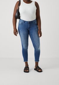 Mörkblå smala jeans med lätt slitning, kombinerade med en vit ärmlös topp och svarta sandaler med remmar. Svart axelväska ingår.