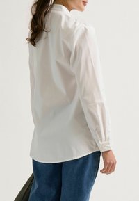 Witte lange mouwen shirt met knoopmanchetten, een gladde textuur en een gebogen zoom. Gecombineerd met blauwe denim broek en een zwarte tas.