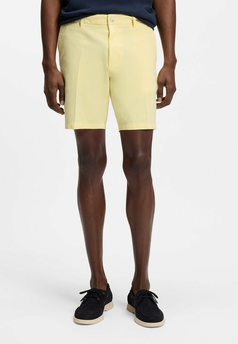 Homme portant un short sur mesure jaune pâle, une chemise bleu marine et des chaussures à lacets noires, debout sur un fond blanc uni.