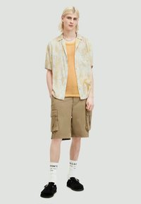 Κοντομάνικο, ανοιχτό κίτρινο πουκάμισο με υφή πάνω από μουσταρδί μπλουζάκι μίας στρώσης; χακί cargo σορτς; μαύρα παπούτσια; λευκές κάλτσες με κείμενο.