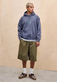 Sweatshirt com capuz em azul ardósia com bolso frontal e gráfico circular, combinado com calções às riscas até ao joelho em verde e castanho, e ténis com padrão.