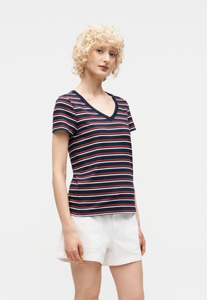 Γυναίκα με κοντά σγουρά ξανθά μαλλιά που φοράει μπλε σκούρη, κόκκινη και λευκή ριγέ μπλούζα με V-λαιμόκοψη και άσπρο σορτς, στέκεται μπροστά σε απλό φόντο.