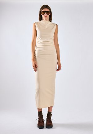 Donna che indossa un vestito midi beige senza maniche, calzini marroni, scarpe marroni stringate e grandi occhiali da sole scuri, in piedi su uno sfondo semplice.