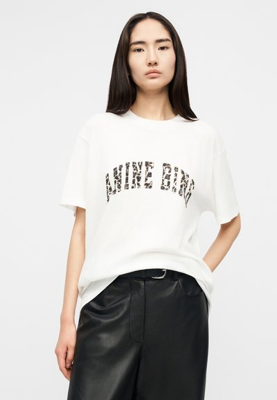 Fiatal nő hosszú sötét hajjal, fehér mintás szöveges pólót és fekete bőrnadrágot viselve, egyszerű háttér előtt áll.