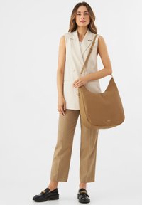 Borsa a tracolla in pelle marrone con una forma morbida e curvata e un dettaglio del logo dorato. Modella indossa un completo beige su misura e mocassini neri.
