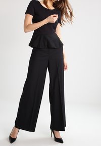 Černé peplum top s texturovanou spodní částí v kombinaci s vysokými kalhotami s širokým nohavicemi. Modelka je obuta v černých vysokých podpatcích. Neutrální pozadí.
