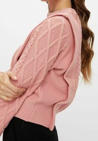 Femme portant un pull en maille torsadée rose pâle avec des motifs texturés, un ourlet côtelé et une coupe ample, associé à un pantalon noir.