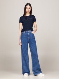 Camiseta de manga corta azul marino con el logotipo "TOMMY JEANS", combinada con jeans de mezclilla azul de pierna ancha y zapatillas blancas, de pie frente a un fondo neutro.