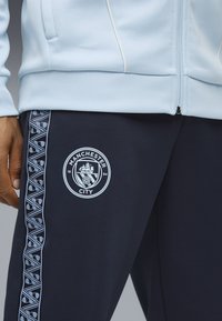 Himmelblaues Zip-Up-Jackett mit geripptem Saum, kombiniert mit dunkelblauen Sweatpants, die ein Manchester-City-Logo und hellblaue gemusterte Seitenstreifen aufweisen.
