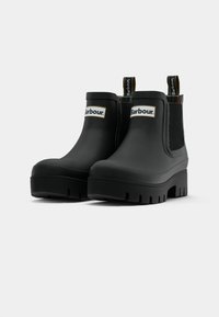 Μαύρες μπότες βροχής Barbour μέχρι τον ασκó με χοντρές σόλες, ελαστικές πλευρές και θηλιές με λογότυπο σε λευκό φόντο.