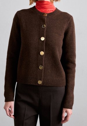 Brązowy kardigan zapinany na guziki, noszony na pomarańczowy golf, zestawiony z ciemnobrązowymi spodniami, na jednolitym tle.