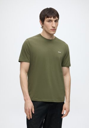Jeune homme portant un t-shirt Hugo vert olive et un pantalon noir, debout devant un fond clair uni avec une expression neutre.