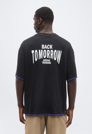 Άνδρας με την πλάτη, φοράει μαύρο T-shirt με λευκή επιγραφή "BACK TOMORROW ADIDAS TRAINING" και μπεζ παντελόνι σε απλό φόντο.