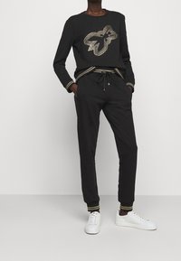 Svart sweatshirt med en texturerad fjärilsdesign i silver, kombinerad med matchande joggers som har guldrandiga muddar och resår i midjan.