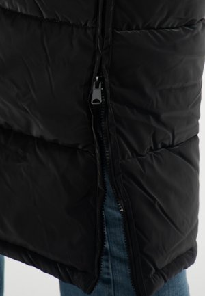 Gilet rembourré noir avec fermeture éclair sur le côté, présentant une texture de tissu lisse et une finition légèrement brillante. Le denim bleu est visible en dessous.