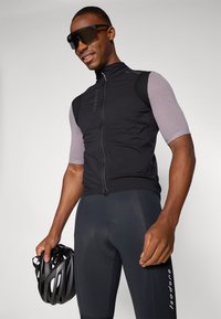 Schwarze Radweste mit kurzärmligem Mesh-Gewebe, kombiniert mit dunklen gepolsterten Shorts. Das Modell hält einen schwarzen Helm. Akzente umfassen ein Logo.