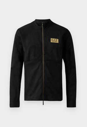 Chaqueta negra de manga larga con cremallera, cuello alto y pequeño parche dorado con el logo de EA7 Emporio Armani en el lado izquierdo del pecho.