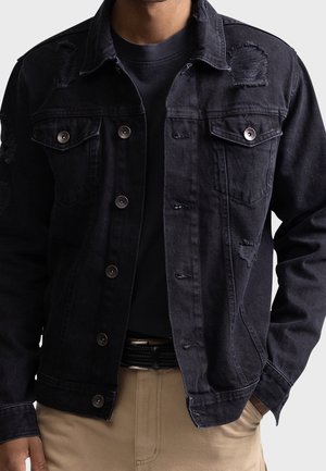 Homme portant une veste en jean noire déchirée par-dessus une chemise noire et un pantalon beige, les mains dans les poches, devant un fond uni.