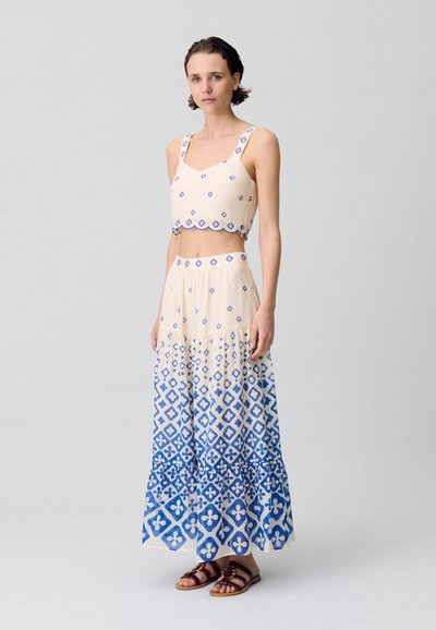 Conjunto de dos piezas que incluye una blusa corta y una falda larga. La tela es ligera, de color crema con patrones geométricos azules. Con dobladillo y tirantes acentuados.