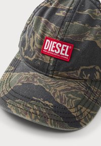 Kamerafarget caps laget av stoff, med en stiv skjerm og et fremtredende rødt broderi-patch med teksten "DIESEL FOR SUCCESSFUL LIVING."