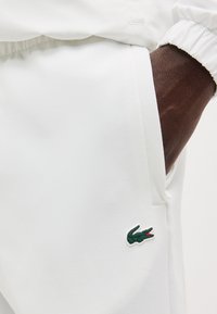 Σκούροχορτο χέρι στην τσέπη του λευκού παντελονιού με ένα μικρό πράσινο λογότυπο κροκοδείλου κοντά στη ραφή της τσέπης.