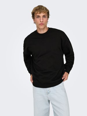 Svart sweatshirt i mjukt tyg med rund halsringning, ribbade muddar och fåll. Ljusblå jeans kompletterar designen. Neutralt bakgrund.