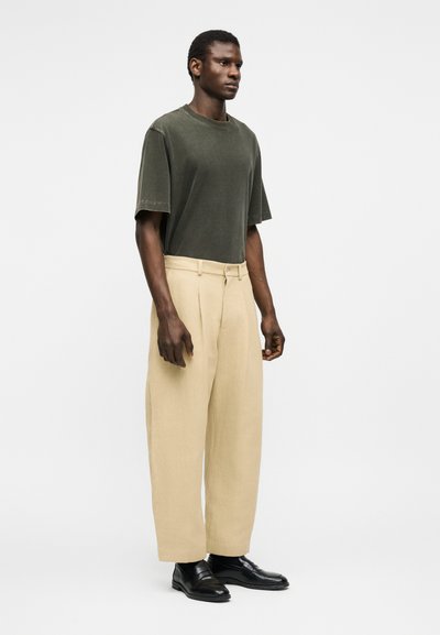 Homme portant une chemise à manches courtes oversize vert foncé, un pantalon large beige et des chaussures de ville noires, debout devant un fond blanc uni.