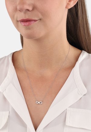 Donna che indossa una collana con simbolo dell'infinito in argento su una sottile catena, abbinata a una blusa bianca a scollatura a V.