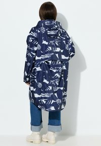 Donna che indossa un impermeabile con cappuccio a fantasia floreale blu navy e bianca, jeans blu corti e stivaletti bianchi, in piedi contro un muro bianco.