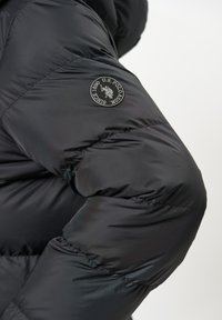 Svart pufferjakke med en glatt tekstur, med et logo-patch på ermet og en quiltet design med avrundet skulderform.