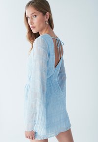 Vaaleansininen mekko, jossa on pitkät, läpinäkyvät, poimutetut hihat ja nauhakiinnitys takana. Mekossa on teksturoitua kangasta ja V-muotoinen selkäosa.
