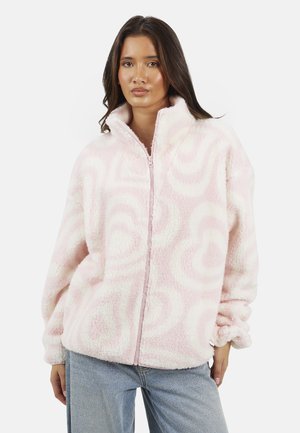 Donna che indossa una giacca in pile rosa e bianca a motivo vorticoso con cerniera frontale e jeans blu, in piedi contro uno sfondo semplice.