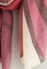 Gewebter Schal mit Streifen in Pink, Creme und Grau, mit weicher Textur und ausgefransten Rändern, vor einem einfarbigen Hintergrund hängend.