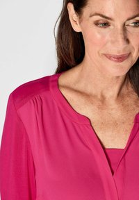 Blusa fucsia con una leggera lucentezza, caratterizzata da un colletto rotondo e spalle plissè, mostrata sul busto di una donna su uno sfondo chiaro.