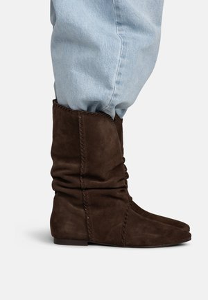 Bottes marron en daim souples mi-mollet avec des détails de coutures, portées avec un jean en denim bleu clair sur un fond uni.