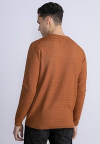 Maglione in cotone arancione bruciato con texture a righe orizzontali, dotato di scollatura a girocollo e maniche lunghe. Indossato sopra jeans scuri.