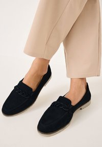 Mustat mokkanahkaiset loafersit, joissa on punottu yksityiskohta avauksen lähellä, kumipohjat ja matala muotoilu, yhdistettynä vaaleanbeigeihin leveisiin housuihin.
