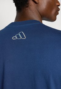 Tumši zilas kokvilnas t-krekls ar mazu baltu izsistā Adidas logotipu aizmugurē, apaļu kakla izgriezumu un īsām piedurknēm.
