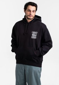 Felpa nera con tasca frontale e cappuccio con cordino. Presenta testo bianco sul petto. Indossata con pantaloni blu-verdi chiari. Design semplice e casual.